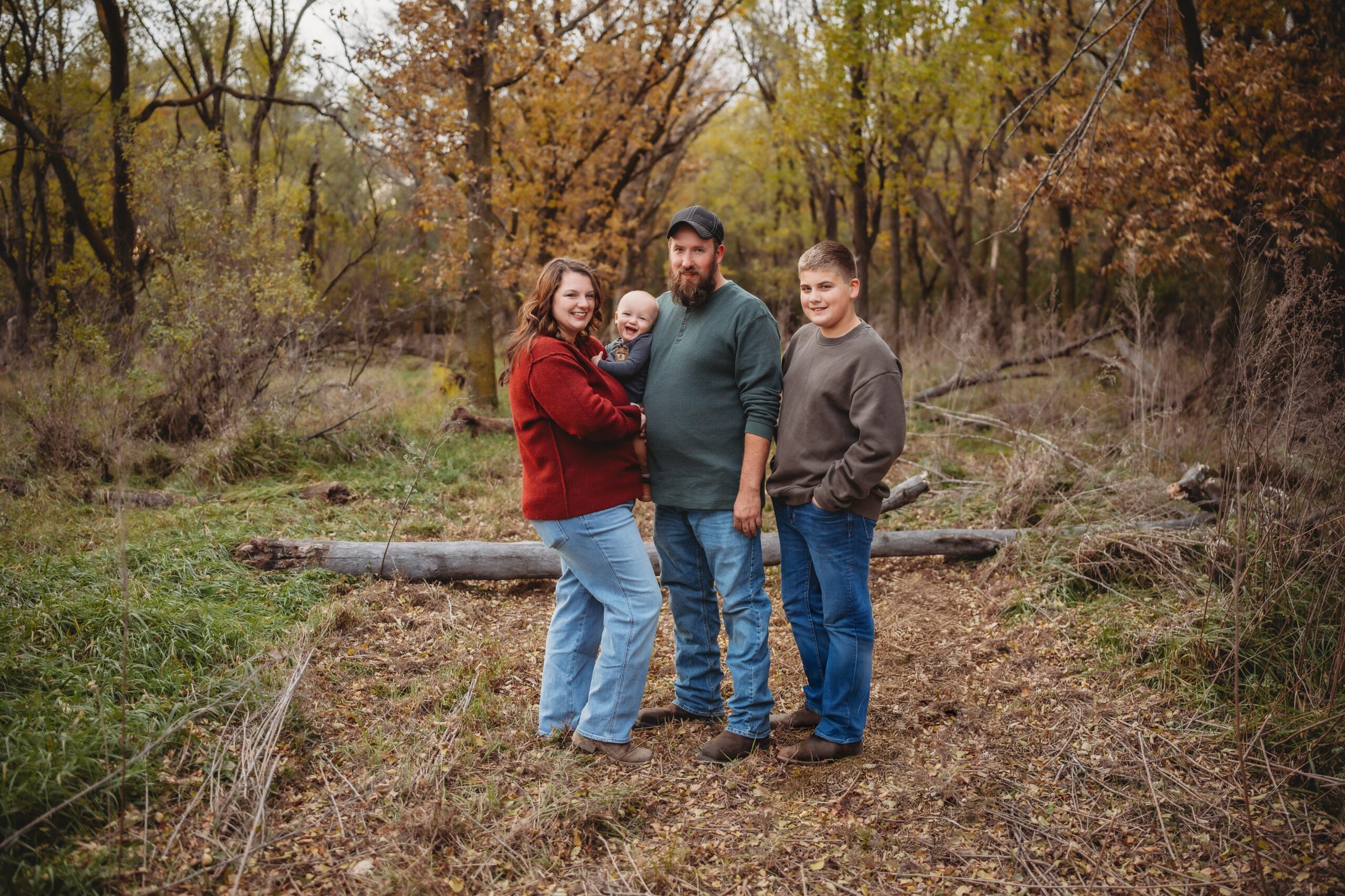 A family of four stands together on a wooded path with autumn trees in the background. The parents hold a baby, and an older child stands beside them.